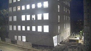 The image shows a five-story concrete building at night, illuminated from within by bright white lights shining through the windows. The construction site around the building is partially visible, with materials and equipment scattered along the ground.