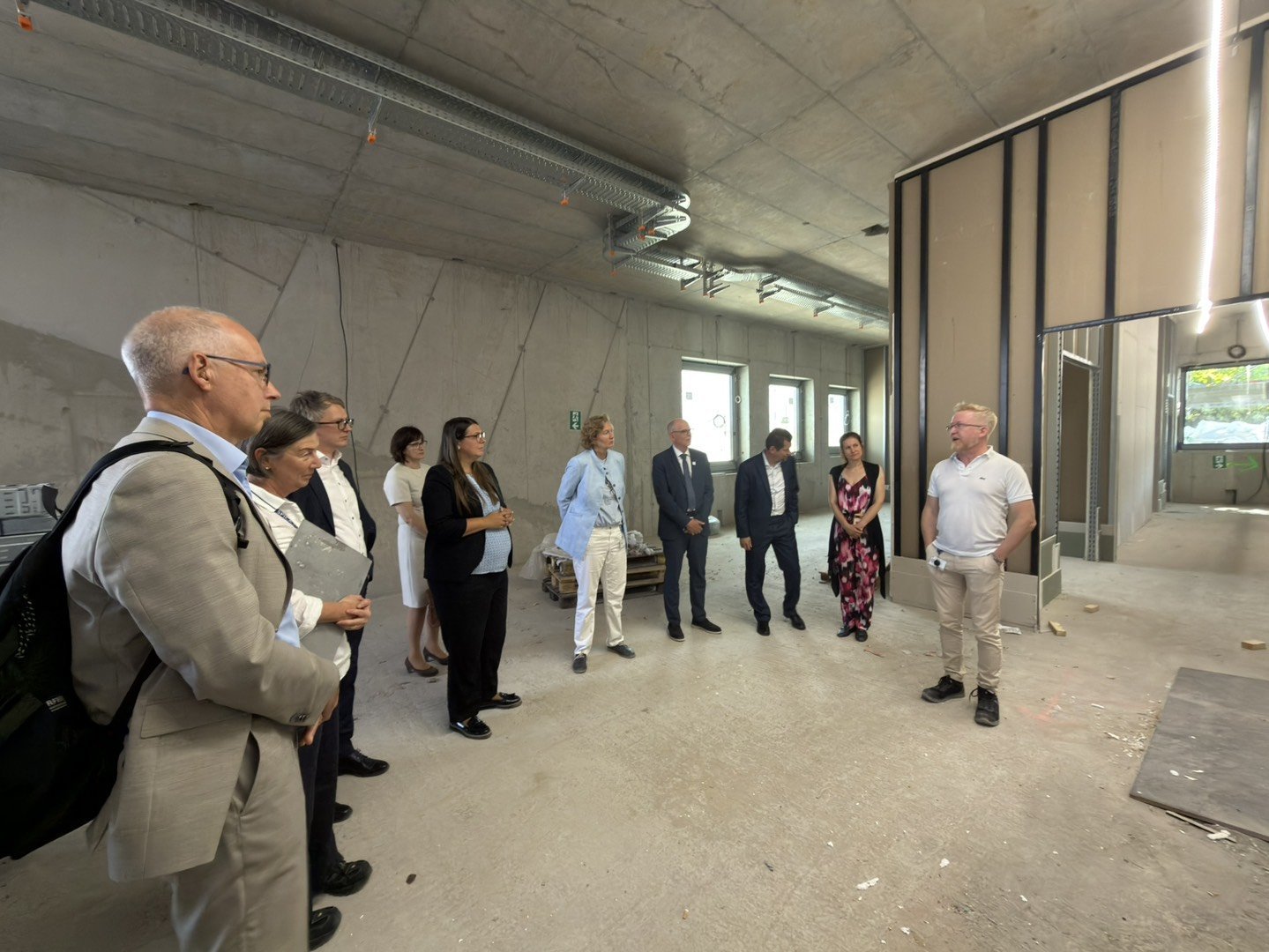 Visitors and hosts stand inside the new building under construction, surrounded by unfinished concrete walls, exposed ceiling infrastructure, and partially completed interior partitions.