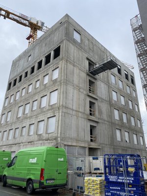 Six-storey concrete building under construction with covered windows, a crane in the background, and an external staircase starting to take shape.