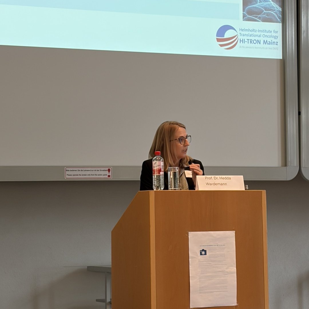 Prof. Dr. Hedda Wardemann speaking at a podium during a scientific conference, with a presentation slide displayed behind her.