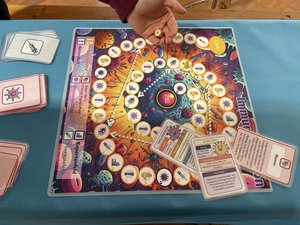 The image shows a colorful educational board game called Immuno Mission, laid out on a turquoise tablecloth. The game board is vibrant, featuring illustrations of cells, viruses, and scientific symbols in a circular path leading to the center marked “ZIEL” (German for “goal”). Along the path are icons representing different categories of questions (guess, action cards, prevention and so on).  A hand is seen holding a small wooden dice above the board, indicating an active game in progress. Game cards with illustrations and German text are spread around the board, including information on immune cells like “T-cells” and “B-cells.” The surrounding area contains decks of pink and blue cards with icon-based illustrations. The game centers arhound the human immune system, aiming to teac players how immune responses work through interactive play.