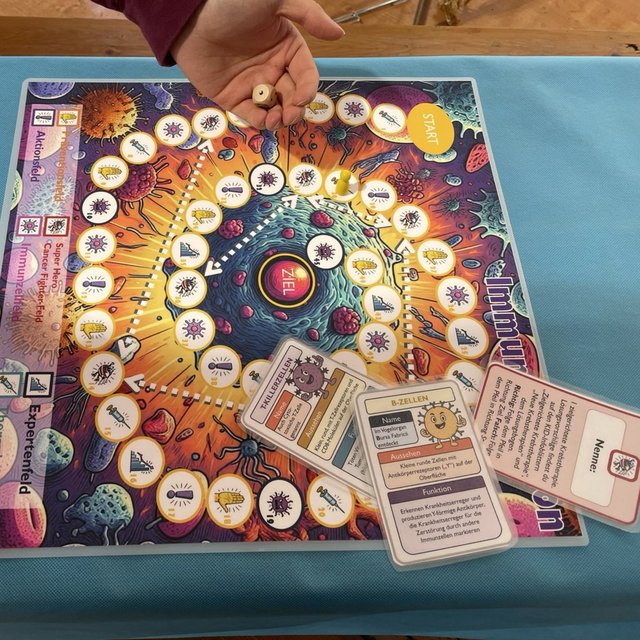 The image shows a colorful educational board game called Immuno Mission, laid out on a turquoise tablecloth. The game board is vibrant, featuring illustrations of cells, viruses, and scientific symbols in a circular path leading to the center marked “ZIEL” (German for “goal”). Along the path are icons representing different categories of questions (guess, action cards, prevention and so on).  A hand is seen holding a small wooden dice above the board, indicating an active game in progress. Game cards with illustrations and German text are spread around the board, including information on immune cells like “T-cells” and “B-cells.” The surrounding area contains decks of pink and blue cards with icon-based illustrations. The game centers arhound the human immune system, aiming to teac players how immune responses work through interactive play.