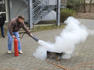 Person using a fire extinguisher to put out smoke from a small training fire device outdoors during a safety exercise.