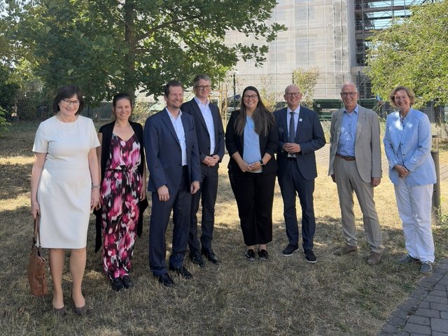 Visitors and hosts stand together outdoors in front of the new HI-TRON Mainz building, which is under construction and partially covered in scaffolding and protective netting.