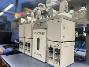 A close-up view of the “Sunscreen” system by Unchained Labs in a laboratory setting. The white analytical instrument is connected with tubes and glowing indicator lights, placed on a lab bench beside glass bottles and a computer monitor.