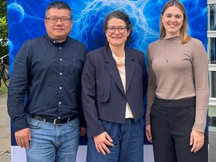 The Platformmembers Claire Conche, Cong Xu and Lea Roth stand in front of a large HI-TRON Mainz banner displaying the institute's name, "Helmholtz Institute for Translational Oncology," above a stylized scientific cell illustration.