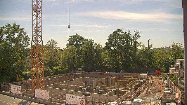 Construction site with crane and foundation work underway, surrounded by trees and clear skies