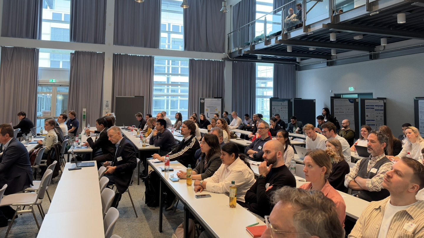 Large audience seated in a modern conference hall, attending a scientific presentation with posters displayed in the background.