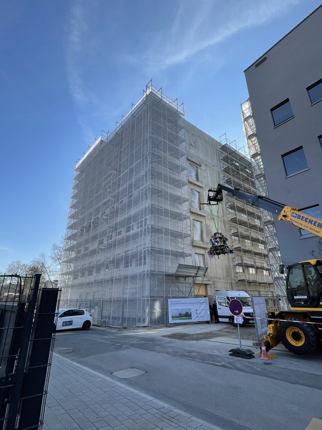 Five-storey building under construction covered in scaffolding, with a crane lifting a traditional topping-out wreath in front of the site.