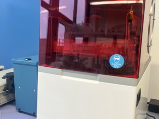 A white desktop 3D printer with a red UV-protective cover, shown operating on a lab bench with its touchscreen displaying a printing progress screen.