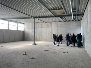 Photo of our site visit: Group of people standing inside a large unfinished concrete room of a building under construction, with exposed walls, ceiling beams, and natural light from high windows.