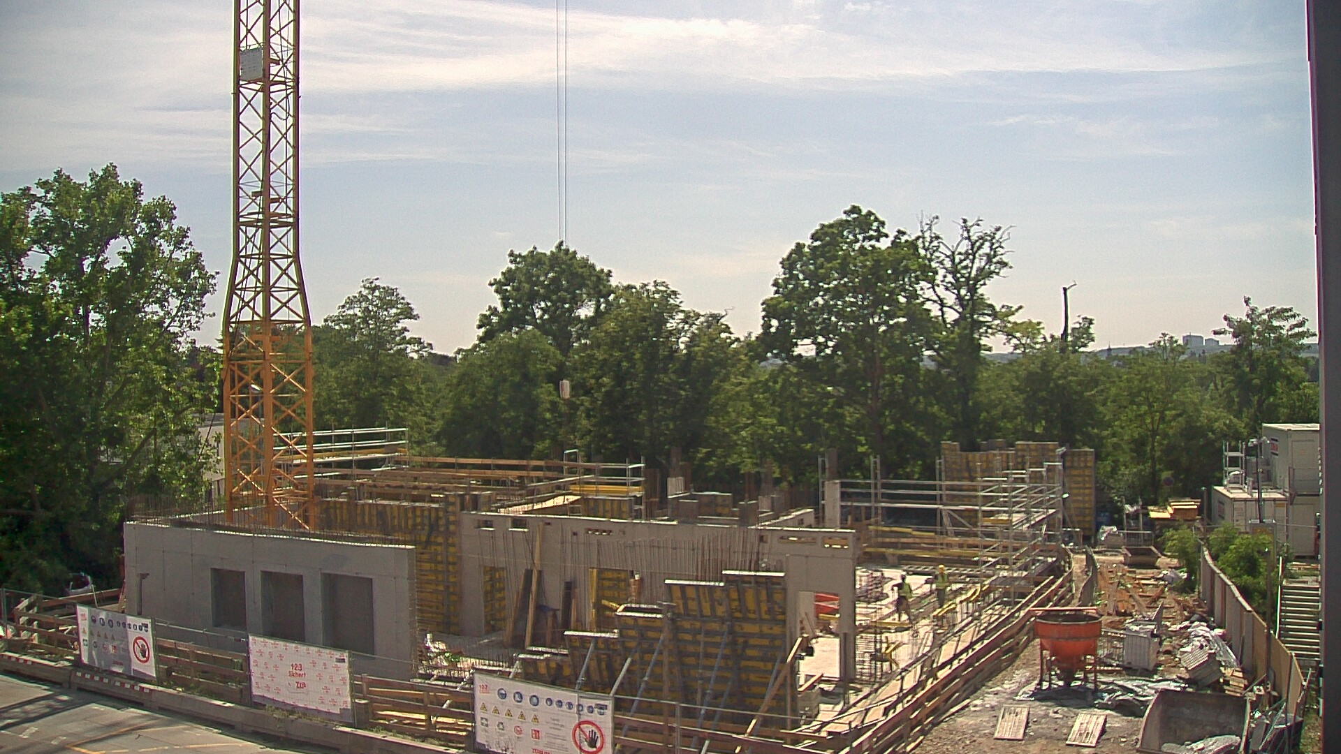 Construction site with partially built concrete walls and yellow formwork structures. A tall yellow crane is visible on the left side, extending vertically out of the frame. Workers are present on the site, surrounded by scaffolding, wooden barriers, and various construction materials. In the background, there are trees under a blue sky with light clouds.
