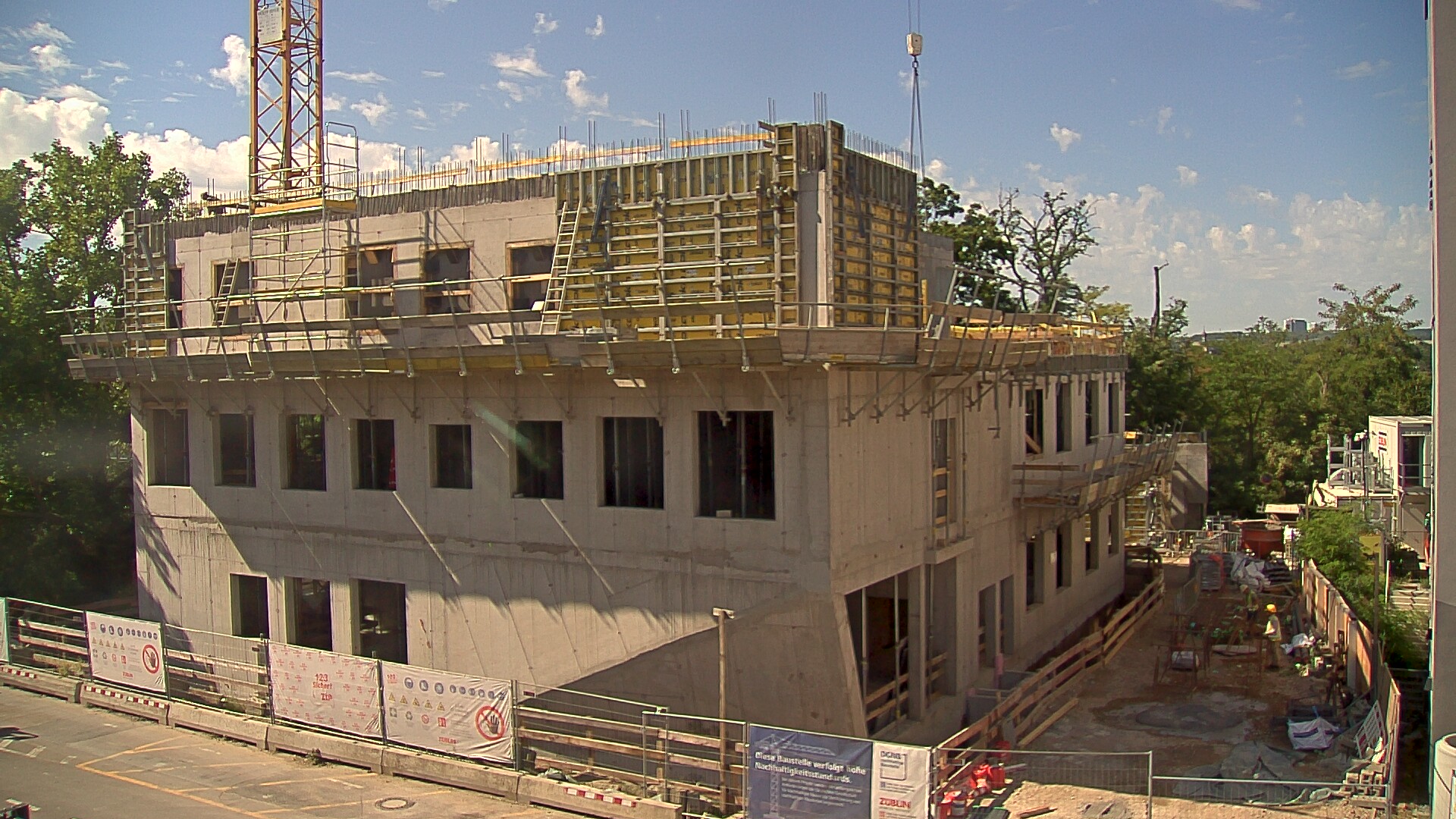 Multi-storey concrete building under construction, with windows installed on lower floors, yellow formwork on the top floor, and a tower crane beside it.