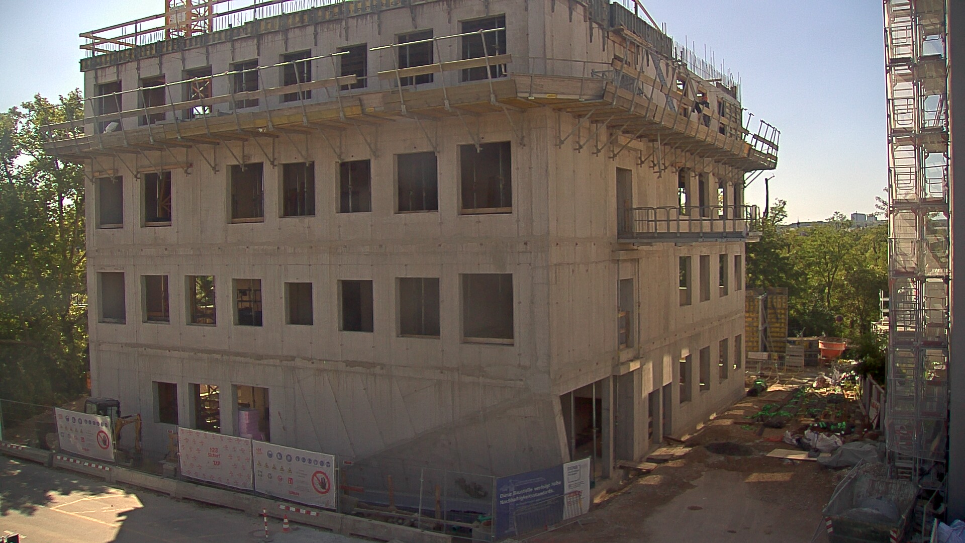 The image shows a five-story concrete building under construction. The facade has open window frames, with scaffolding and safety railings installed on the upper floors. In front of the building, construction materials and a small excavator are visible.