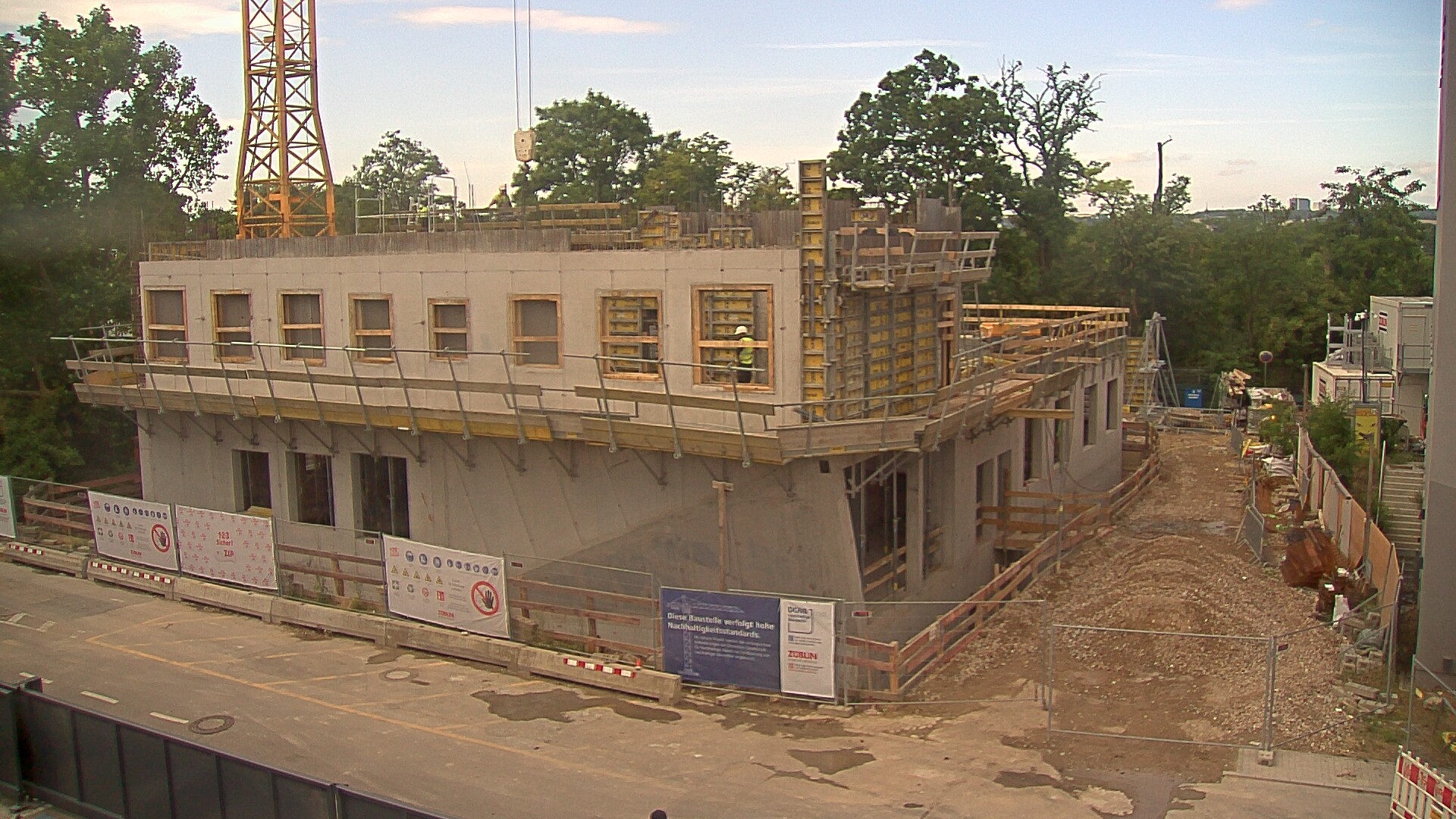A two-story concrete building under construction, with scaffolding and safety railings along the upper floor, a crane in the background, and a gravel pathway with construction materials and temporary site cabins on the right side.