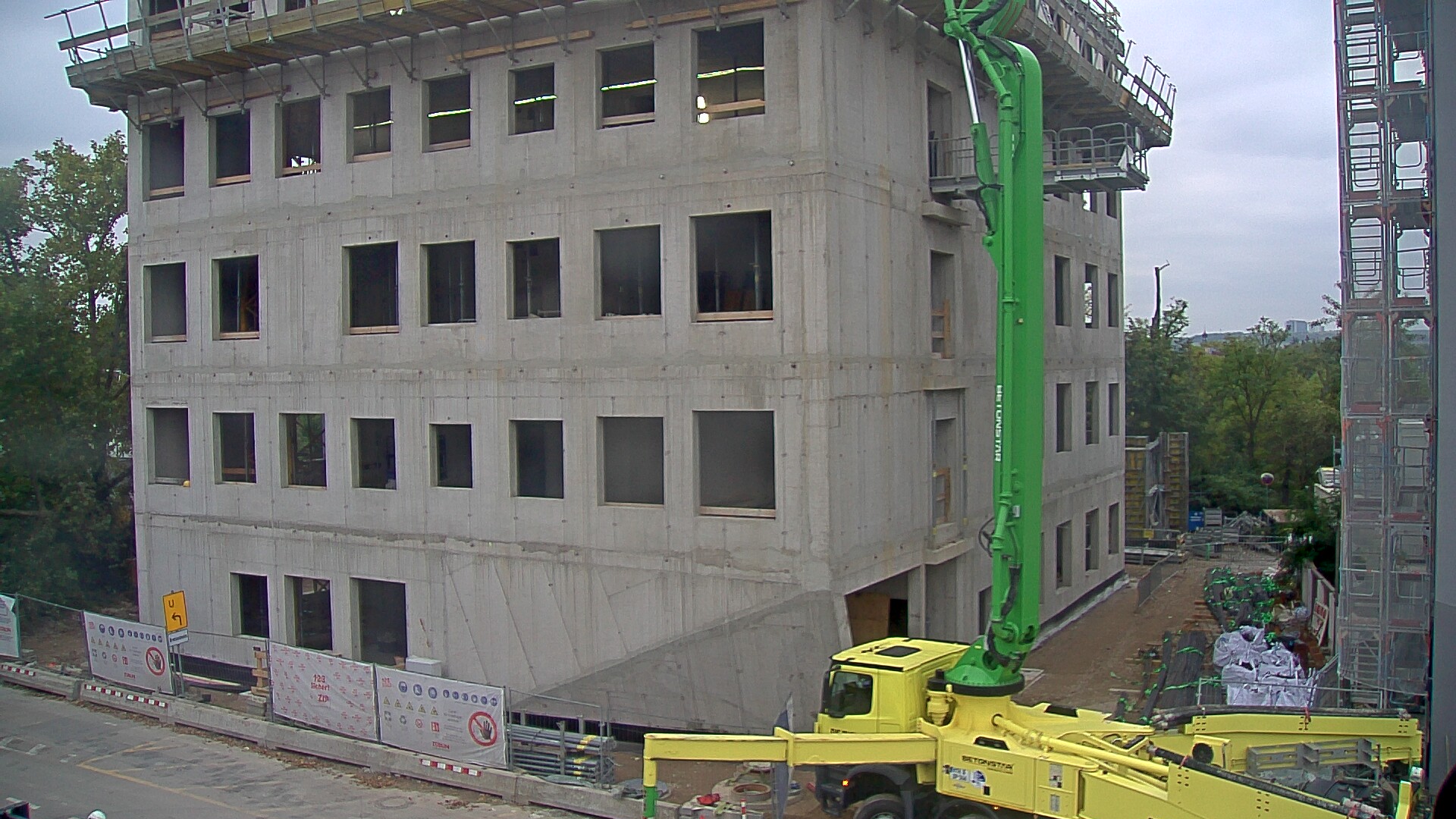 The image shows a six-story concrete building under construction. A bright green concrete pump truck is positioned next to the structure, with its long arm extended toward the upper floors. The windows are still open, scaffolding is visible at the top, and construction materials are scattered around the fenced site.