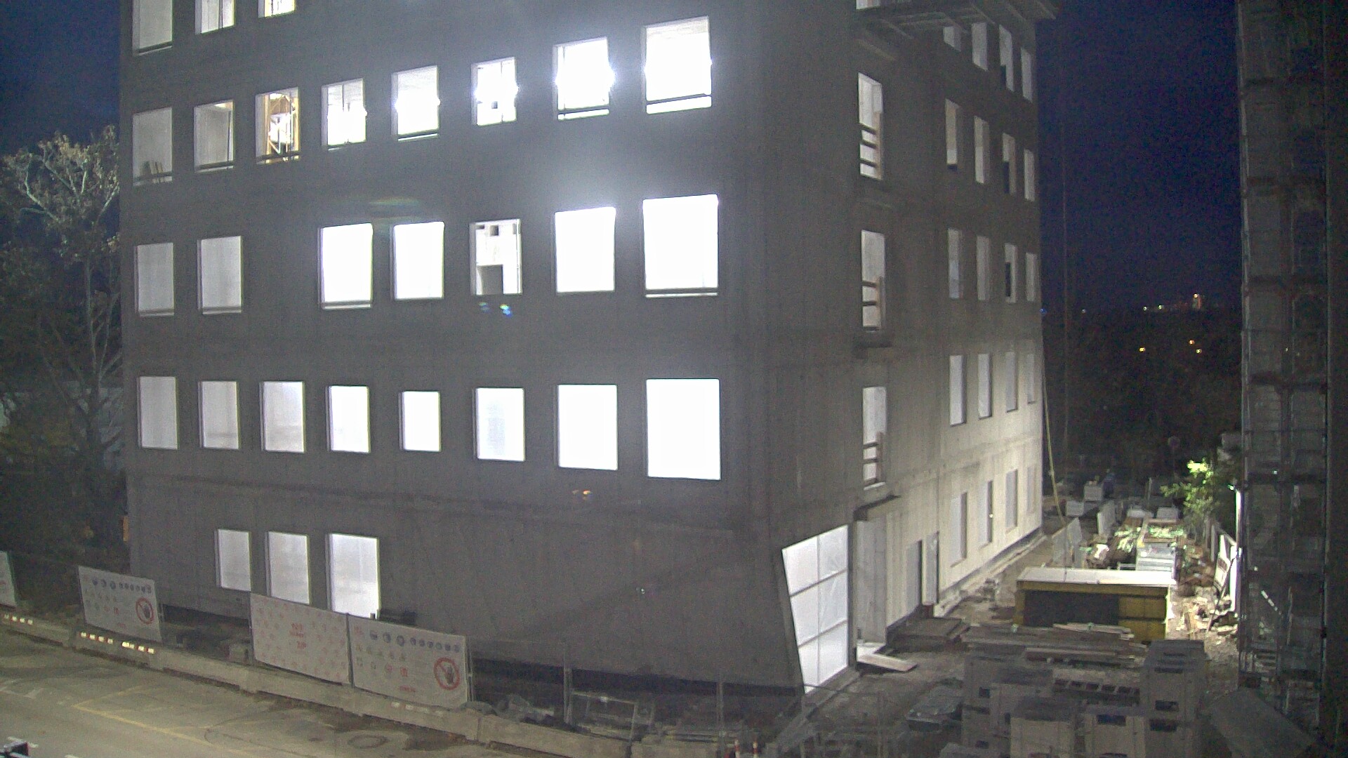 The image shows a five-story concrete building at night, illuminated from within by bright white lights shining through the windows. The construction site around the building is partially visible, with materials and equipment scattered along the ground.