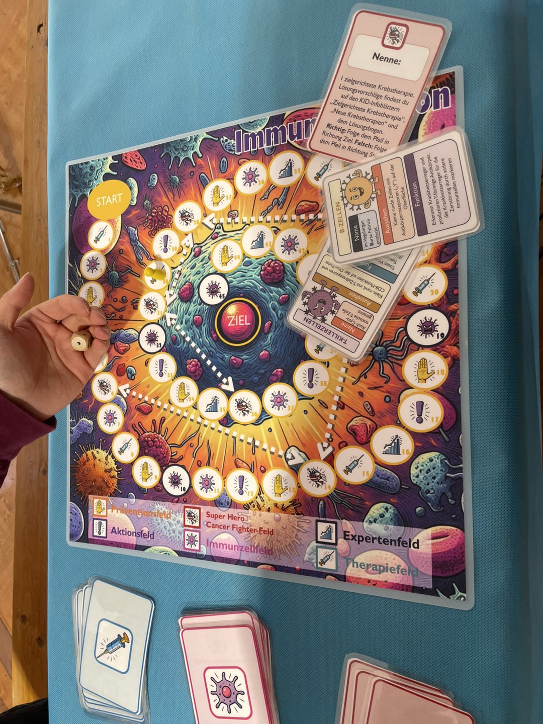 The image shows a colorful educational board game called Immuno Mission, laid out on a turquoise tablecloth. The game board is vibrant, featuring illustrations of cells, viruses, and scientific symbols in a circular path leading to the center marked “ZIEL” (German for “goal”). Along the path are icons representing different categories of questions (guess, action cards, prevention and so on).  A hand is seen holding a small wooden dice above the board, indicating an active game in progress. Game cards with illustrations and German text are spread around the board, including information on immune cells like “T-cells” and “B-cells.” The surrounding area contains decks of pink and blue cards with icon-based illustrations. The game centers arhound the human immune system, aiming to teac players how immune responses work through interactive play.