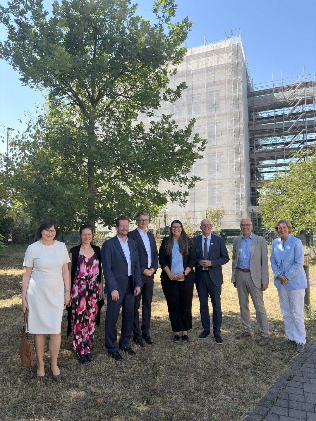 Visitors and hosts stand together outdoors in front of the new HI-TRON Mainz building, which is under construction and partially covered in scaffolding and protective netting.