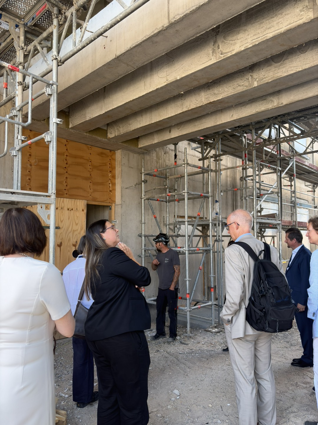Visitors and hosts tour the new building under construction, standing beneath reinforced concrete floors and surrounded by scaffolding while receiving an explanation of the construction progress.