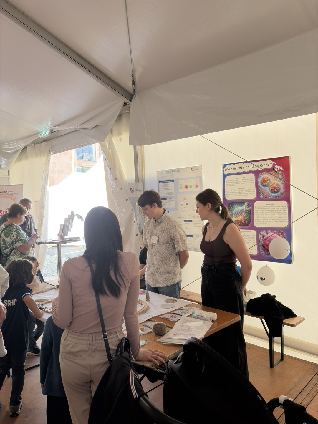 Visitors interact with staff at an educational booth inside a tent, featuring scientific posters about cancer research on the wall.