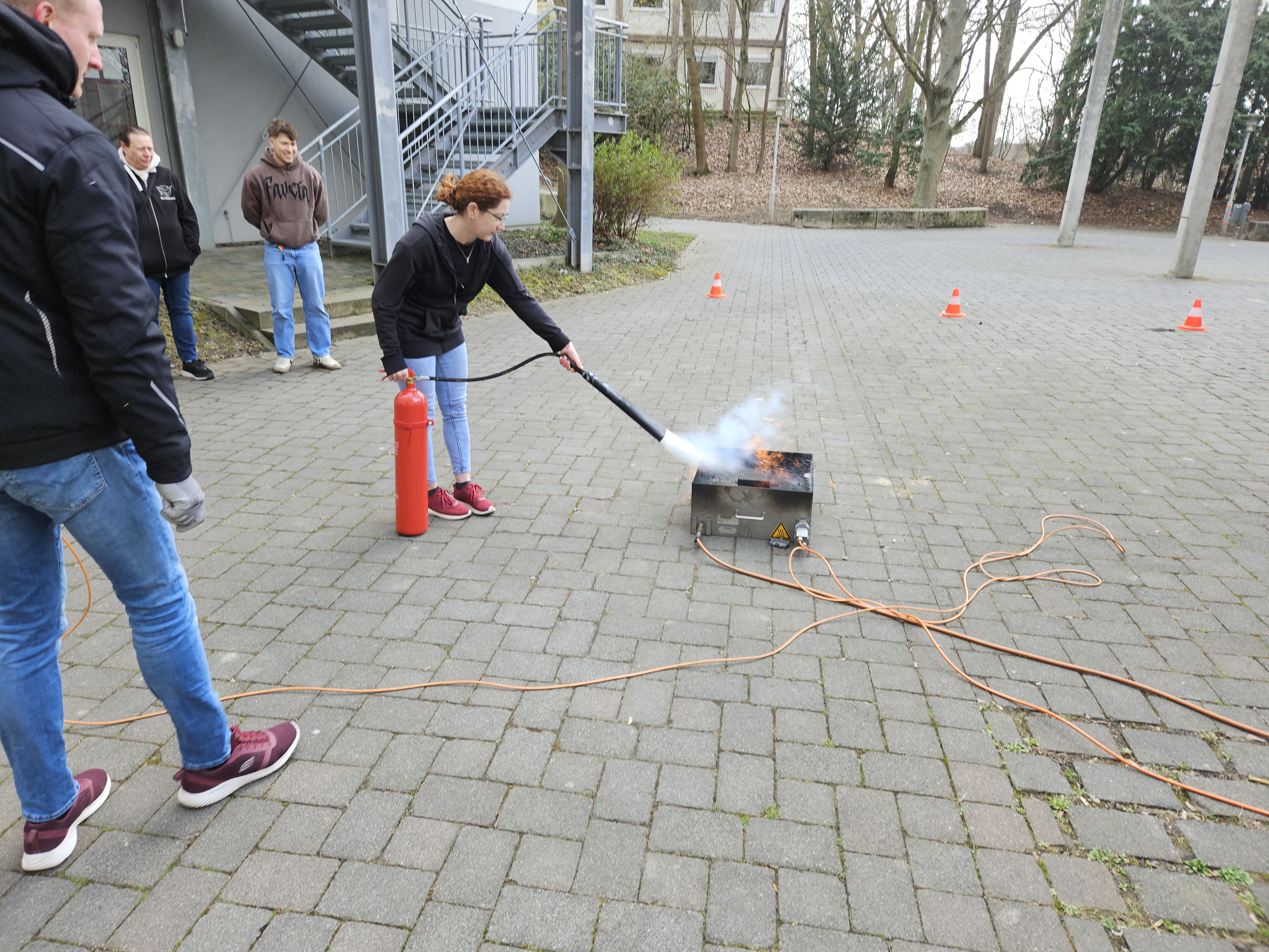 Person using a fire extinguisher to put out smoke from a small training fire device outdoors during a safety exercise.