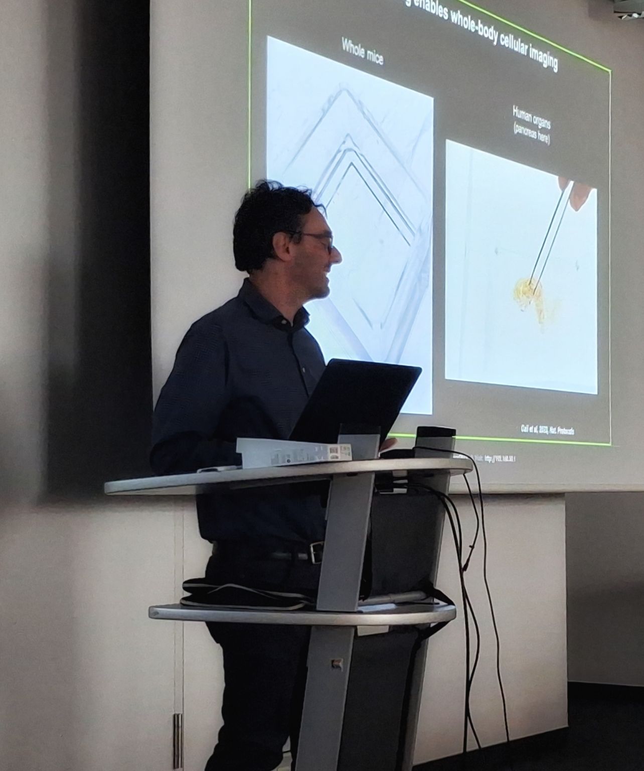 Markus Elsner stands at a lectern giving a scientific presentation, with a projected slide on cellular imaging in the background.