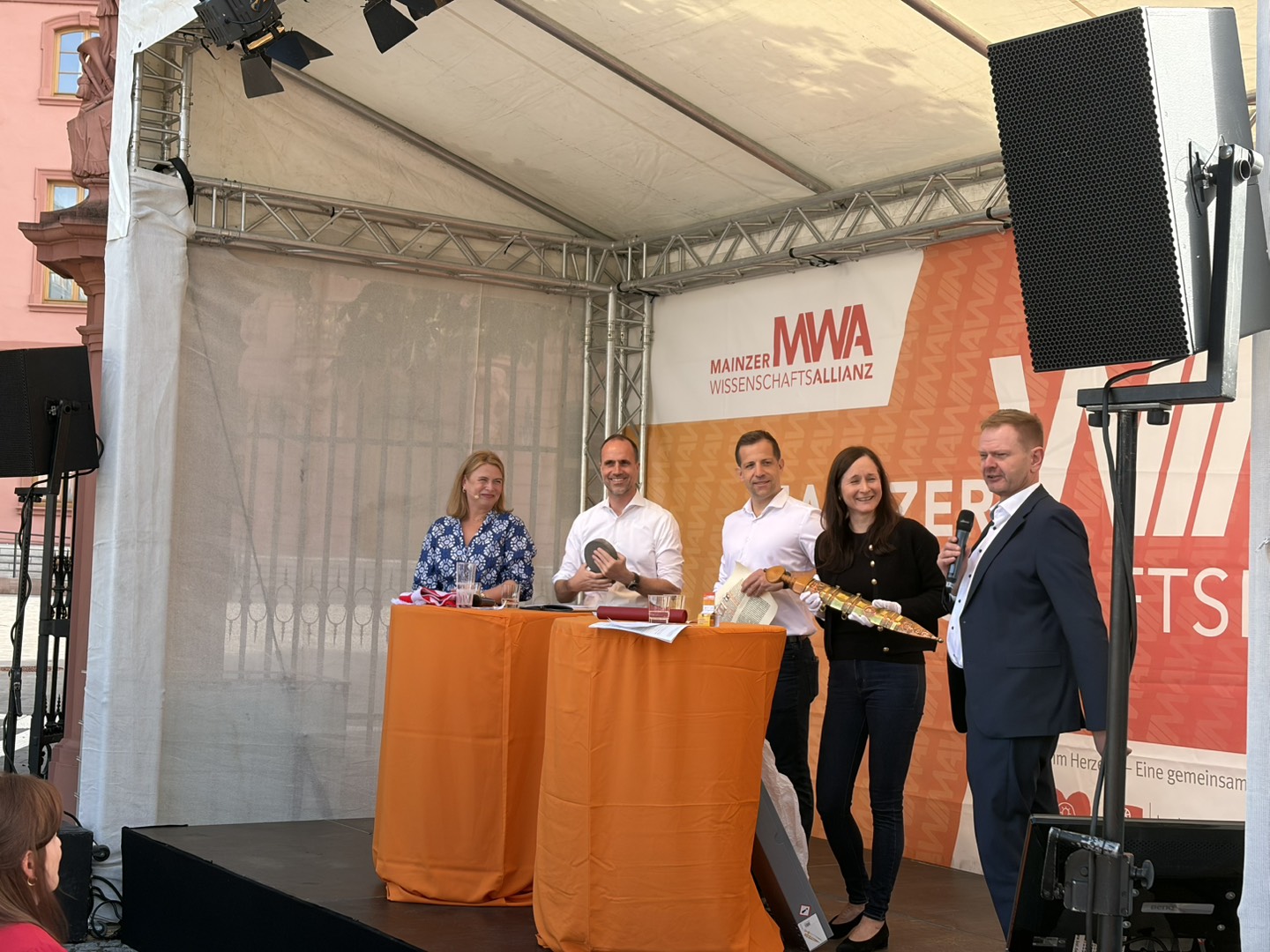 A group of five people on a stage under a orange banner with"MWA Mainzer Wissenschaftsallianz written on it. There is a speaker with a microphone and others holding ceremonial items (a sword, a printed page and a millstone).