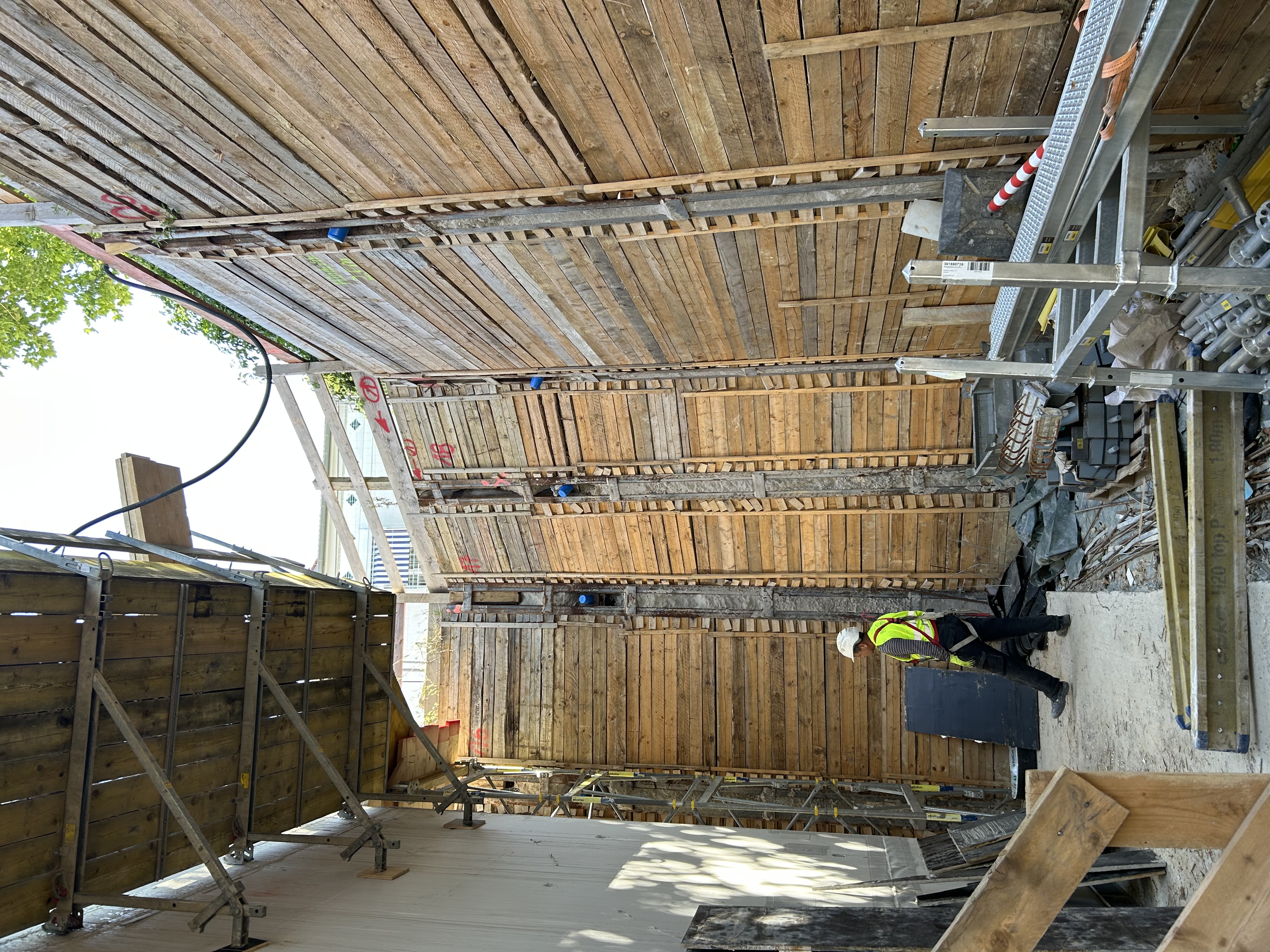A construction worker in high-visibility gear stands inside a deep excavation site, surrounded by tall timber retaining walls with scattered construction materials on the ground.