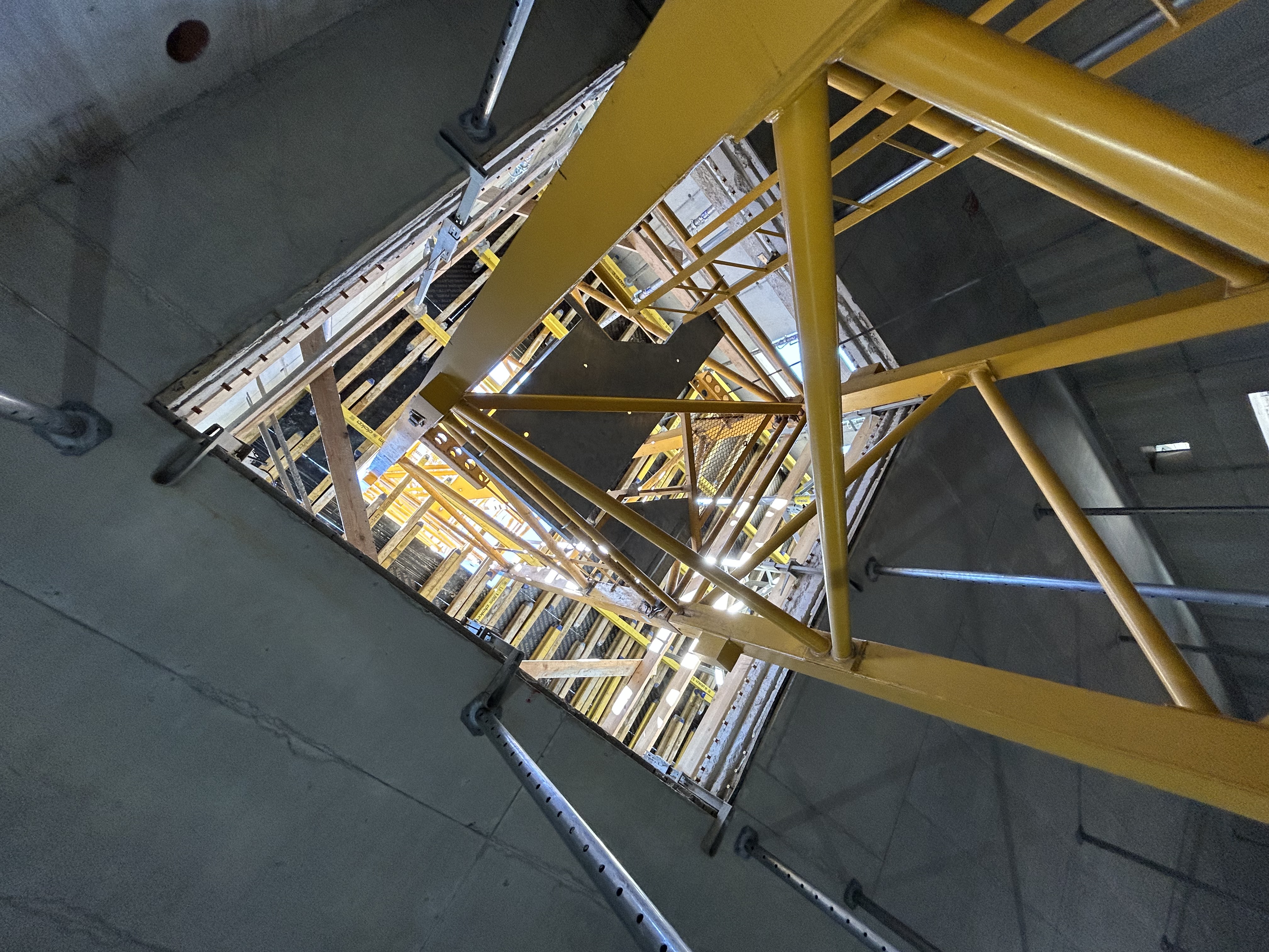 View from below looking up through a concrete shaft, revealing a yellow steel structure framed by formwork and scaffolding with daylight streaming from above.