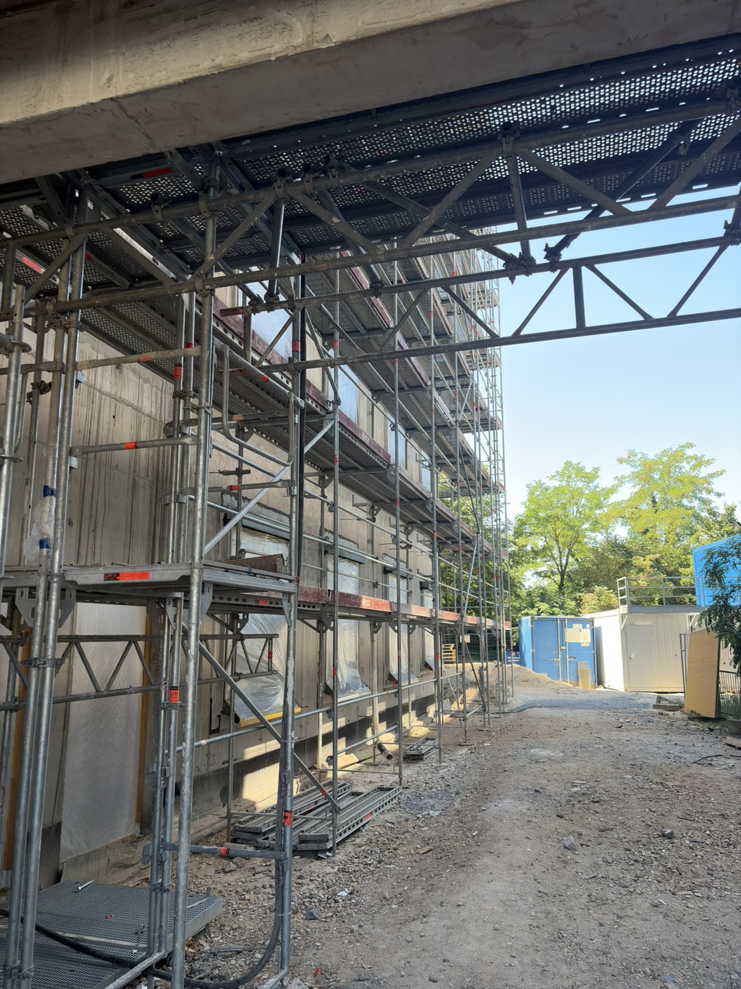 Scaffolding surrounds the exterior of the new building under construction, with unfinished concrete walls, covered window openings, and a temporary construction path alongside the building.