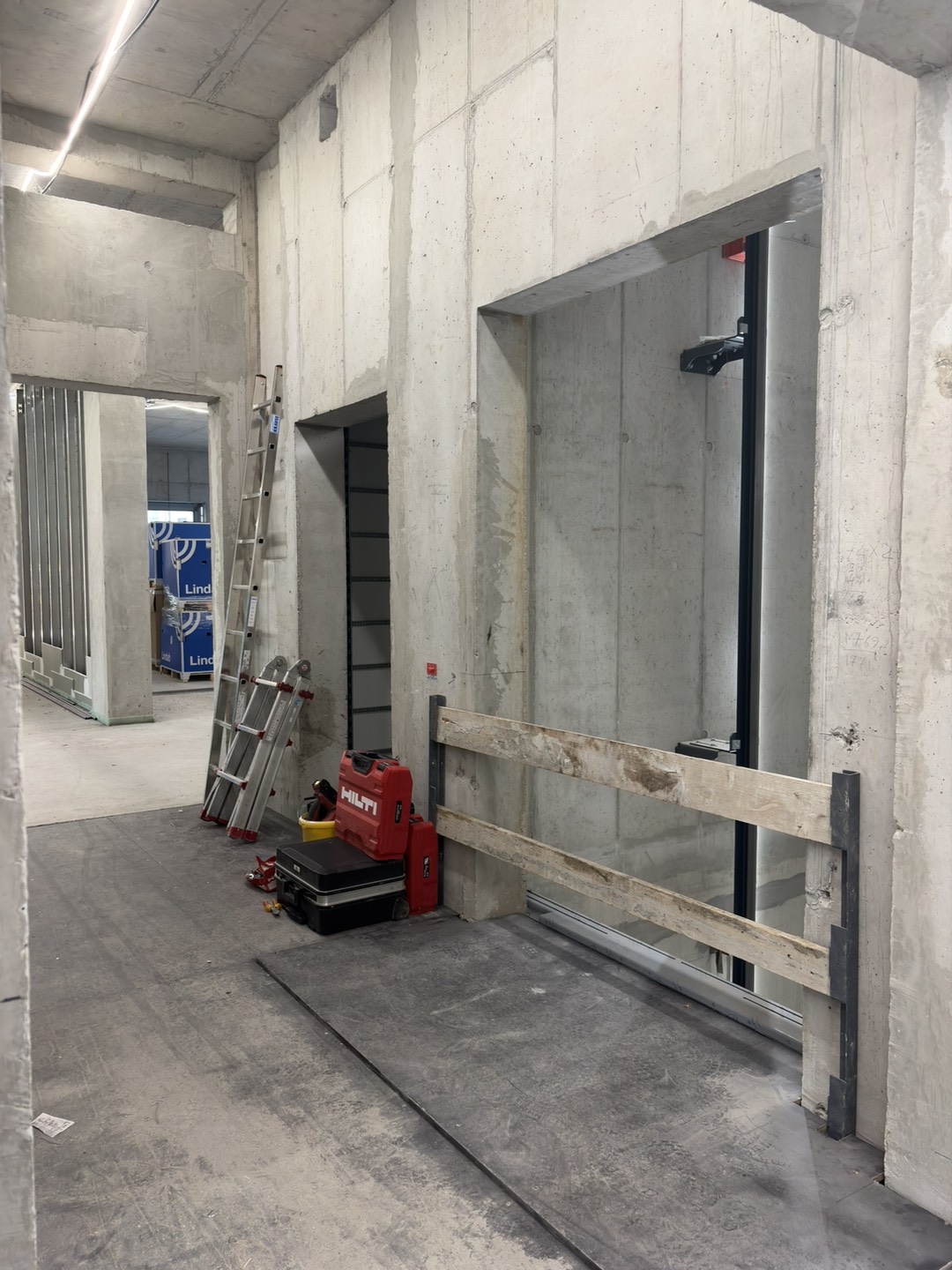 Unfinished interior area of the new building with exposed concrete walls, construction equipment, and an open elevator shaft secured by a temporary wooden barrier.