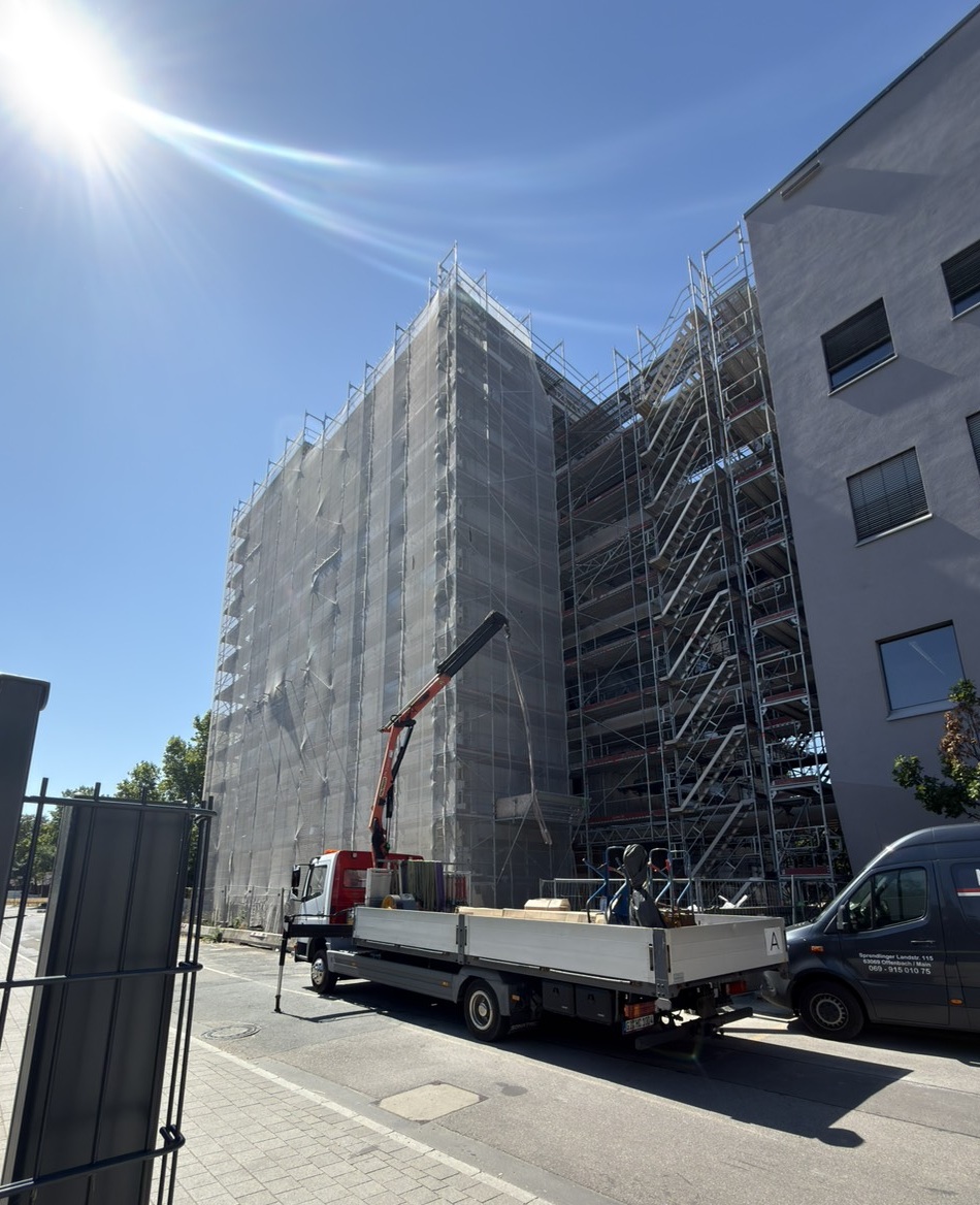The new multi-storey HI-TRON Mainz building is surrounded by scaffolding and protective netting, while connecting bridges between the new building and the adjacent building are under construction.