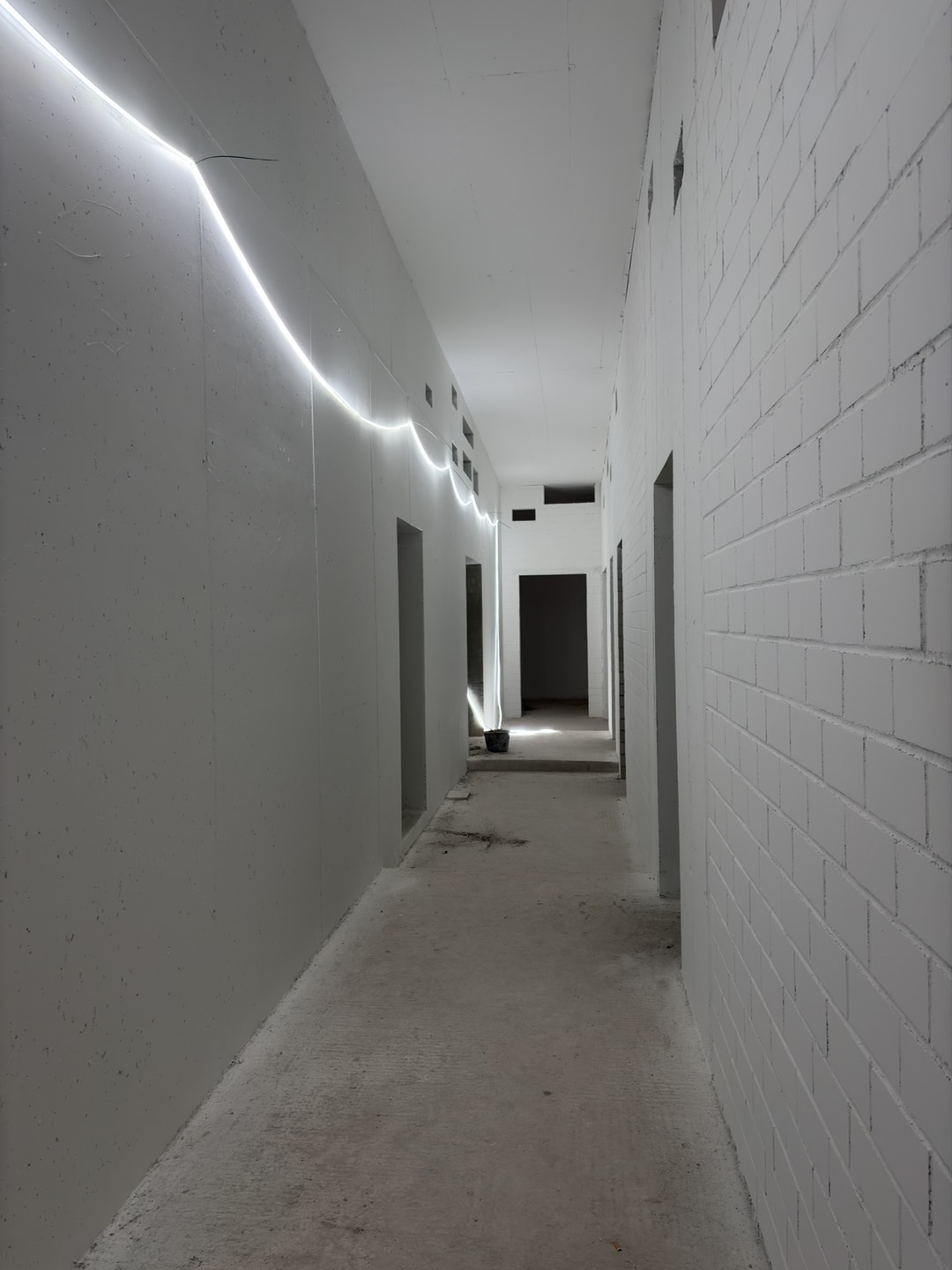 An unfinished corridor inside the new building, with white concrete and brick walls, open doorways, a bare concrete floor, and temporary lighting along the wall.