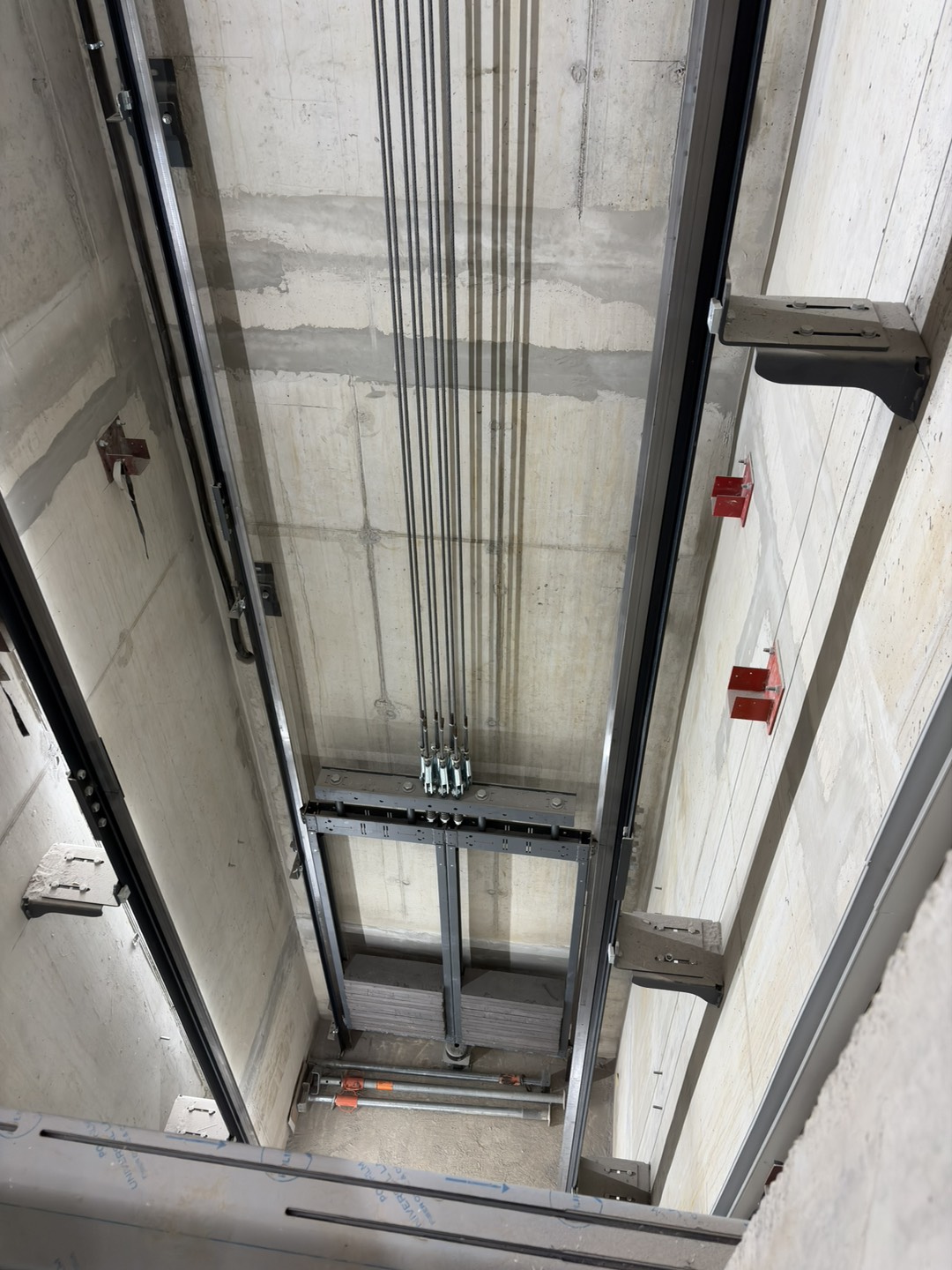 View down an unfinished elevator shaft, showing exposed guide rails, suspension cables, mounting brackets, and concrete walls during installation.