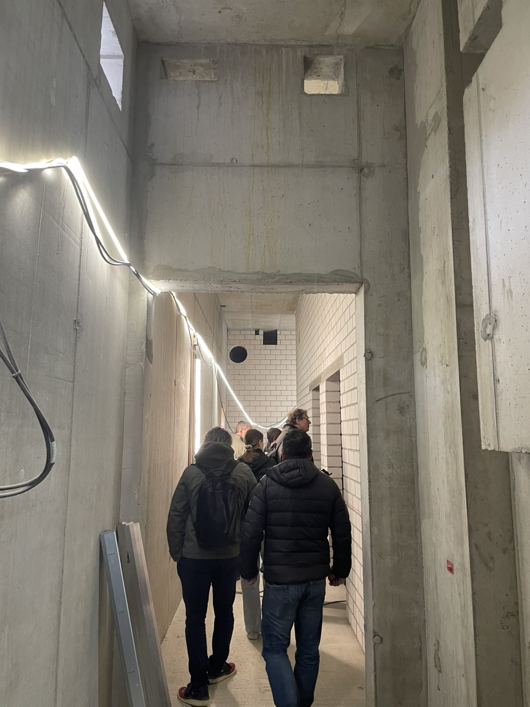 Photo of our site visit: Group of people walking through a narrow unfinished corridor with exposed concrete walls, temporary lighting, and partially installed tiled sections.