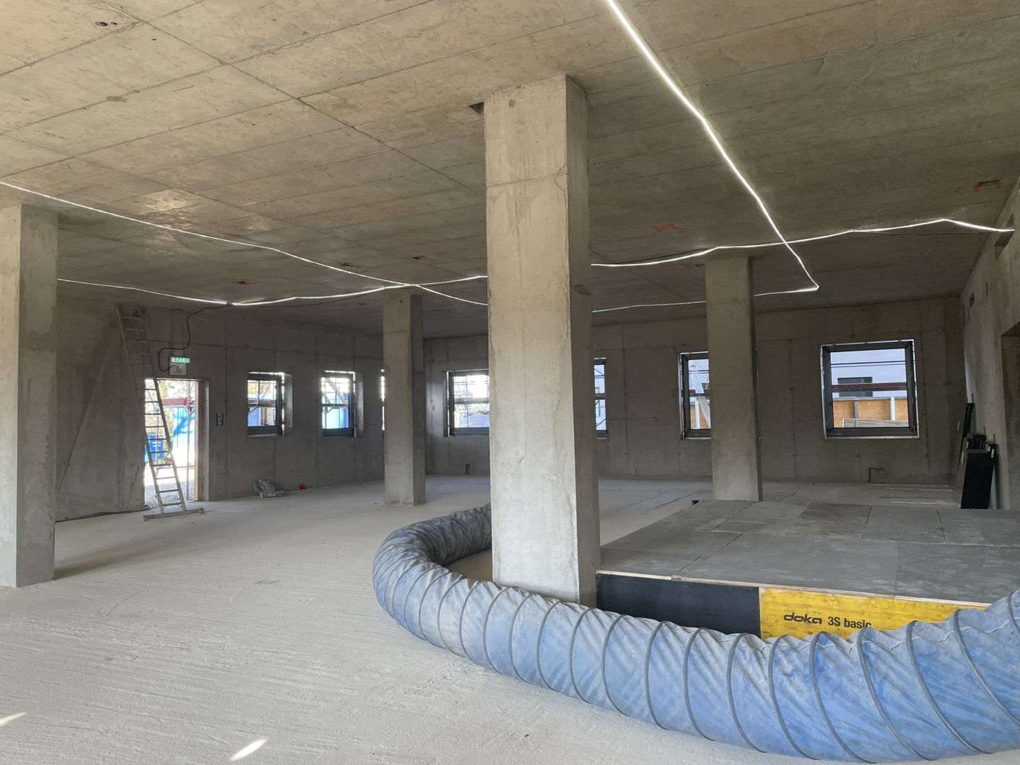 Photo of our site visit:Large unfinished interior space with exposed concrete columns, multiple windows, temporary lighting cables on the ceiling, and construction equipment on the floor.