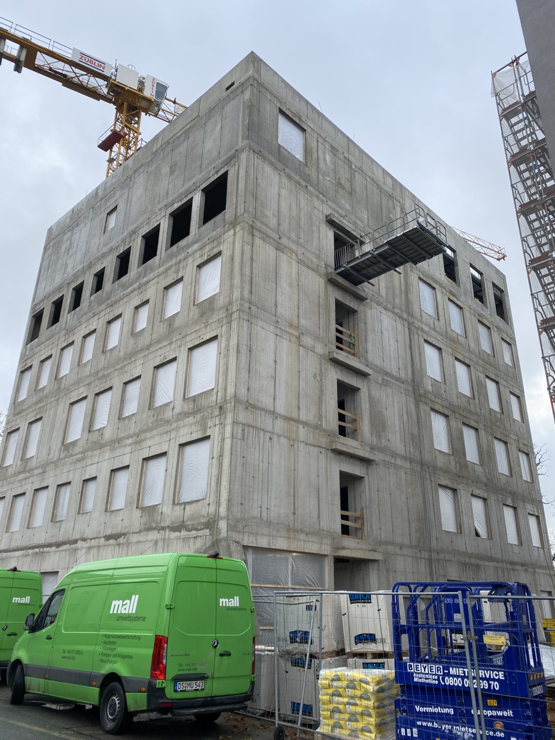 Six-storey concrete building under construction with covered windows, a crane in the background, and an external staircase starting to take shape.